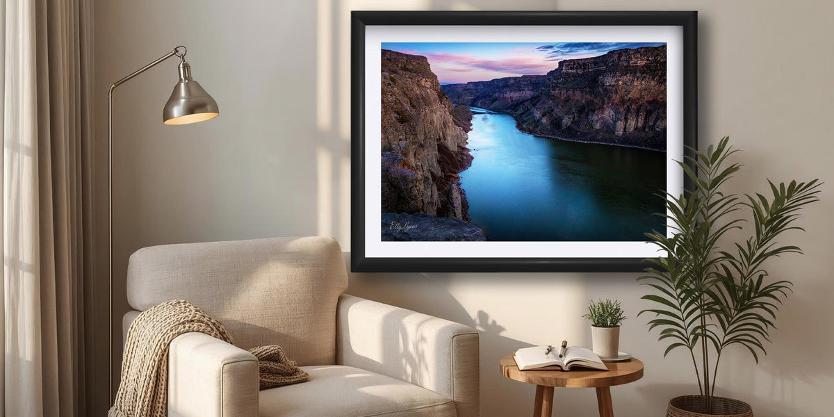 An framed image of the Snake River Canyon at sunrise sits above a reading chair with a lamp overhead next to a window with soft morning light coming in.