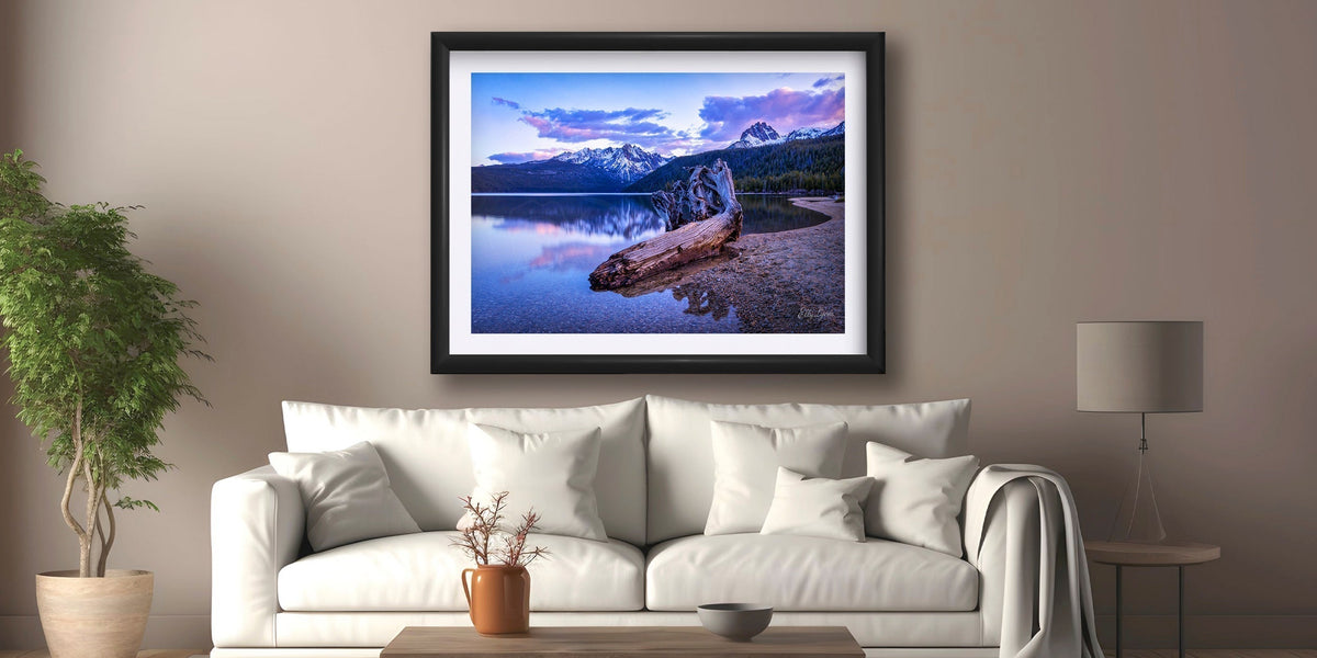 A framed image of Redfish Lake at sunset with the snow capped Sawtooth Mountains in the background hangs above a neutral couch in a cozy and comfortable living room setting with soft light filtering in through an adjacent window.
