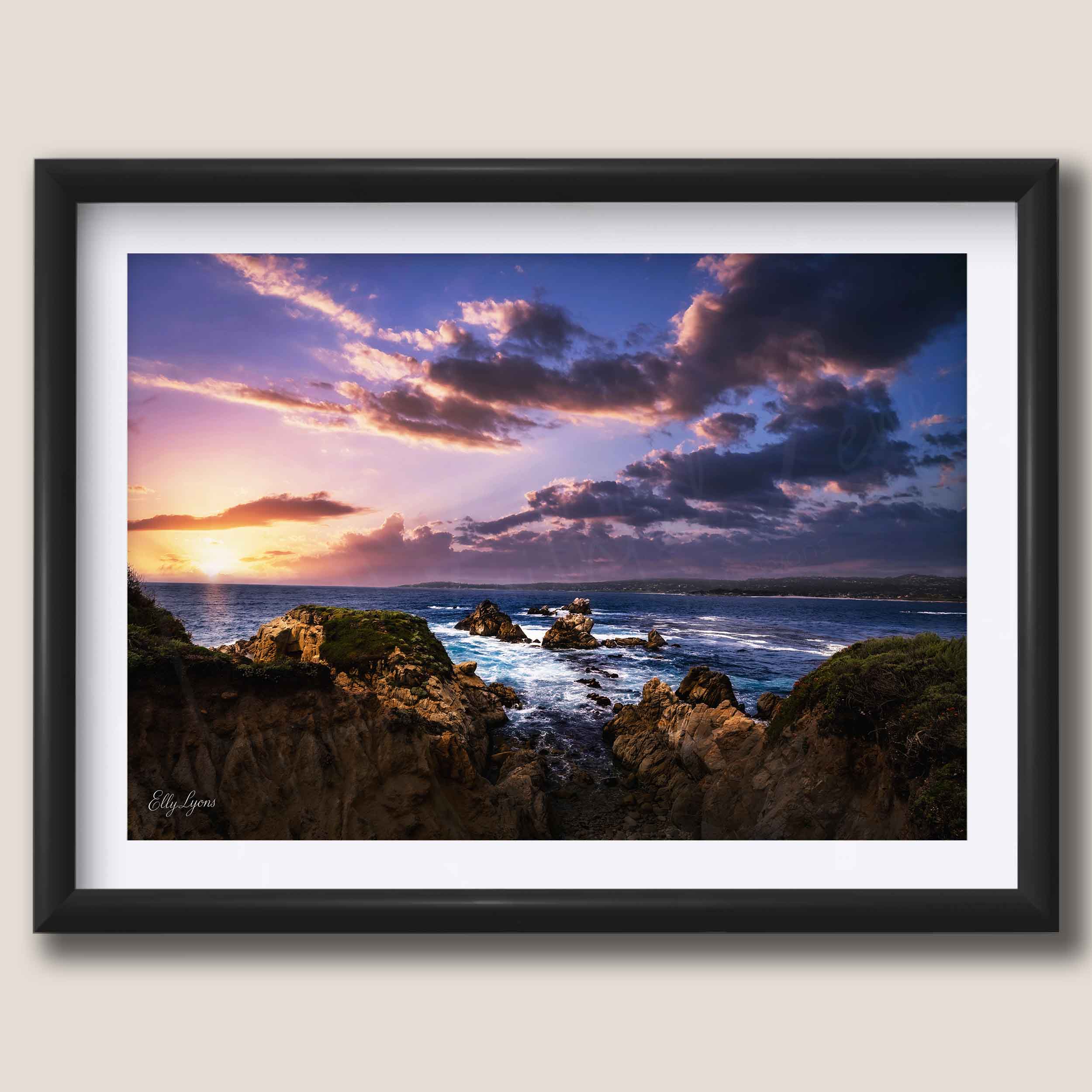 A framed coastal landscape photograph showing rocky cliffs, ocean waves, and dramatic sunset clouds in purple, pink, orange, and blue tones, presented in a black frame with a white mat against a light beige background.